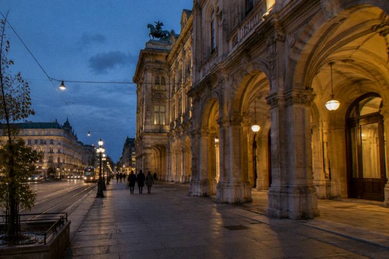 Dusk under the stone arcades of the Vienna State Opera, warm light on the arches and small silhouettes walking away.