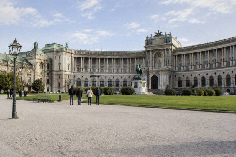 The curved colonnaded facade of the Neue Burg wing of the Hofburg across Heldenplatz, a small group of visitors seen from behind.