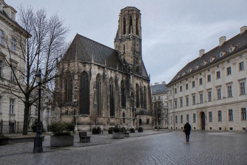 The weathered Gothic stone facade of the Minoritenkirche on Minoritenplatz, with a lone passer-by walking away.