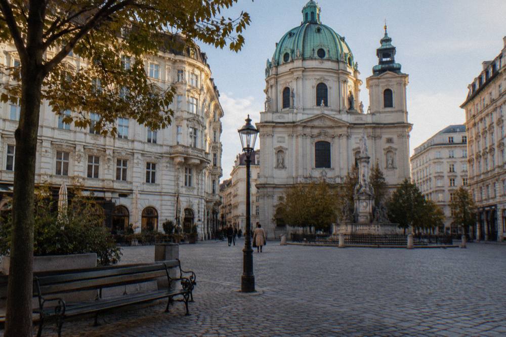 A cobbled central Vienna square with Baroque facades in late afternoon light and empty bench seats