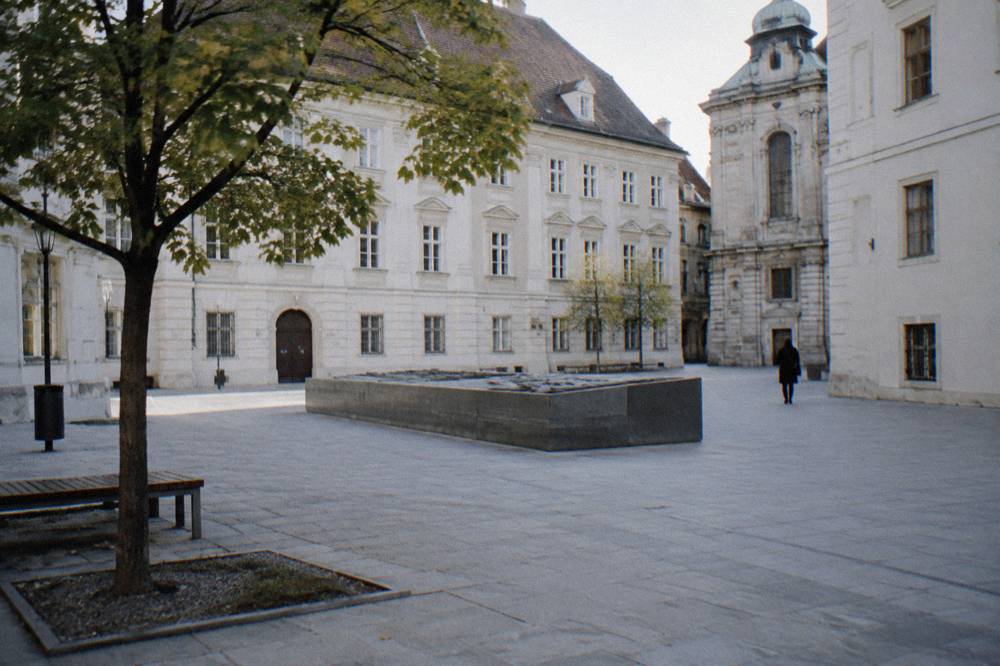 The Judenplatz square in central Vienna with its plain pale facades, a low block memorial at the centre and a distant walker