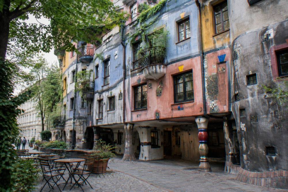 The tiled colourful courtyard-side buildings of the Hundertwasser Village across the street, with empty cafe tables outside