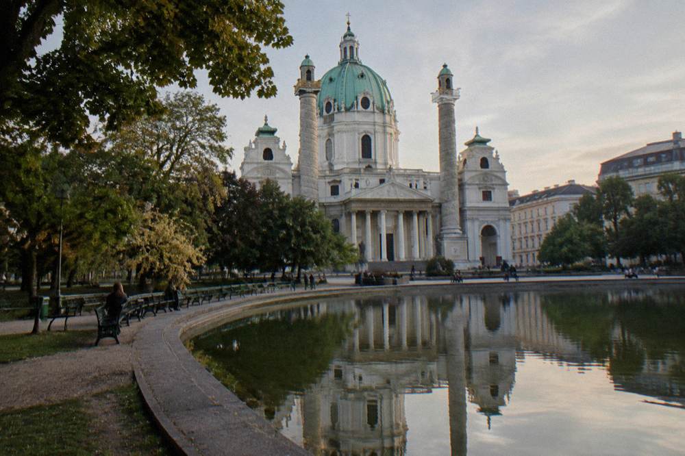 The Karlsplatz pond reflecting the Karlskirche facade with park benches and trees along the water's edge