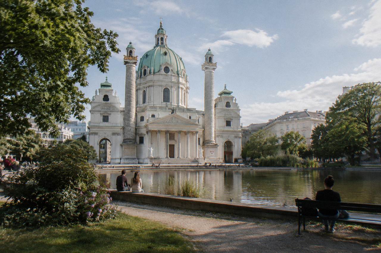 The Karlskirche green dome and twin columns behind the reflecting pond in Resselpark under a bright sky