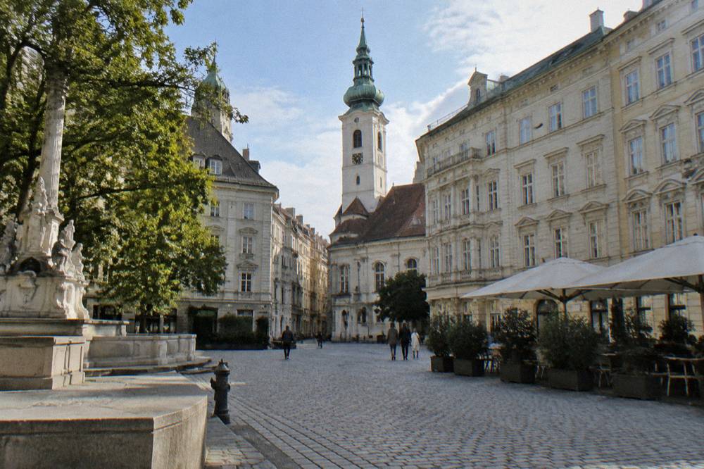 A cobbled square in central Vienna with a church tower rising behind the rooftops in afternoon light