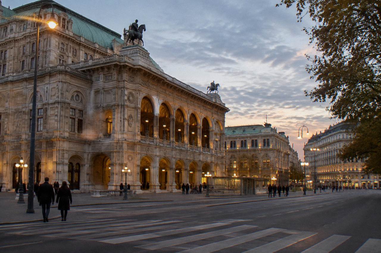 The State Opera facade and adjoining Opernring under an evening sky, arcades lit warm and a pedestrian crossing in front