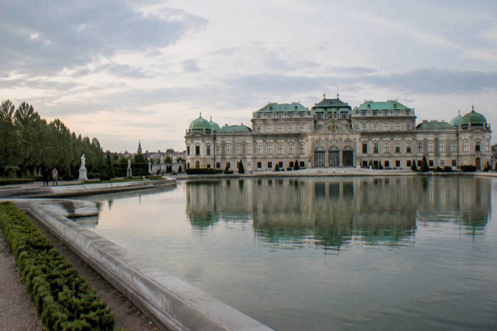 The Upper Belvedere facade seen across the long reflecting pond with its green copper roofs, photographed from the water's edge in soft light