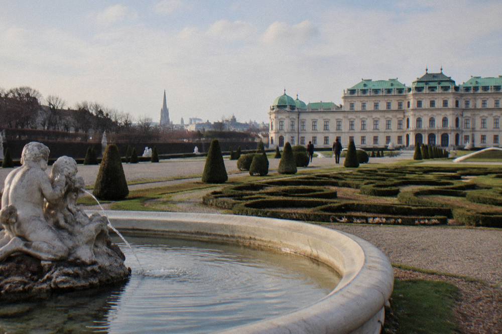 A stone basin fountain and a clipped parterre in the Belvedere garden with the Upper Belvedere rooftop in the background