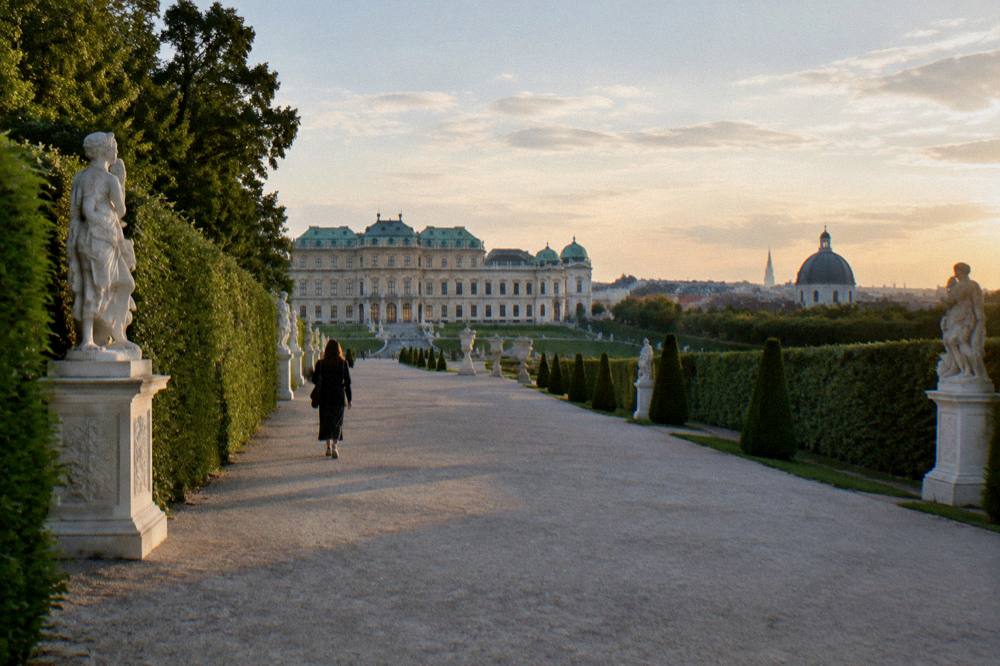 A wide gravel path between tall trimmed hedges and stone statues in the Belvedere garden, seen from behind a distant walker in the late afternoon