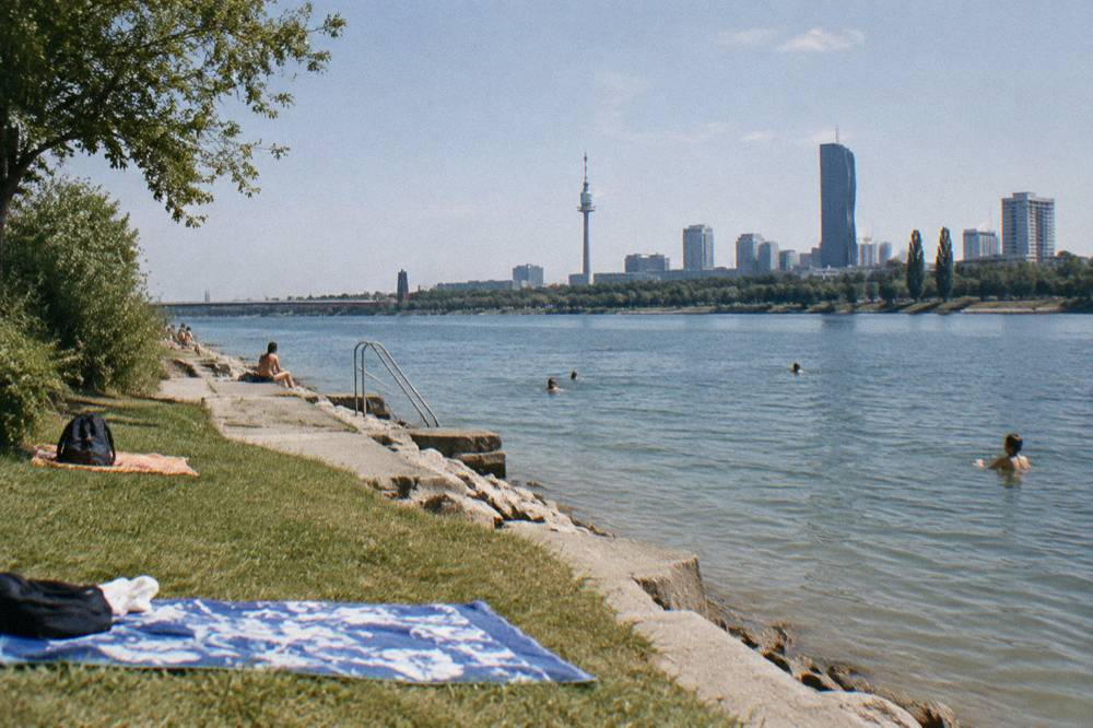 A concrete and stone bathing shore on the Donauinsel with towels on the grass and a few distant swimmers in the water