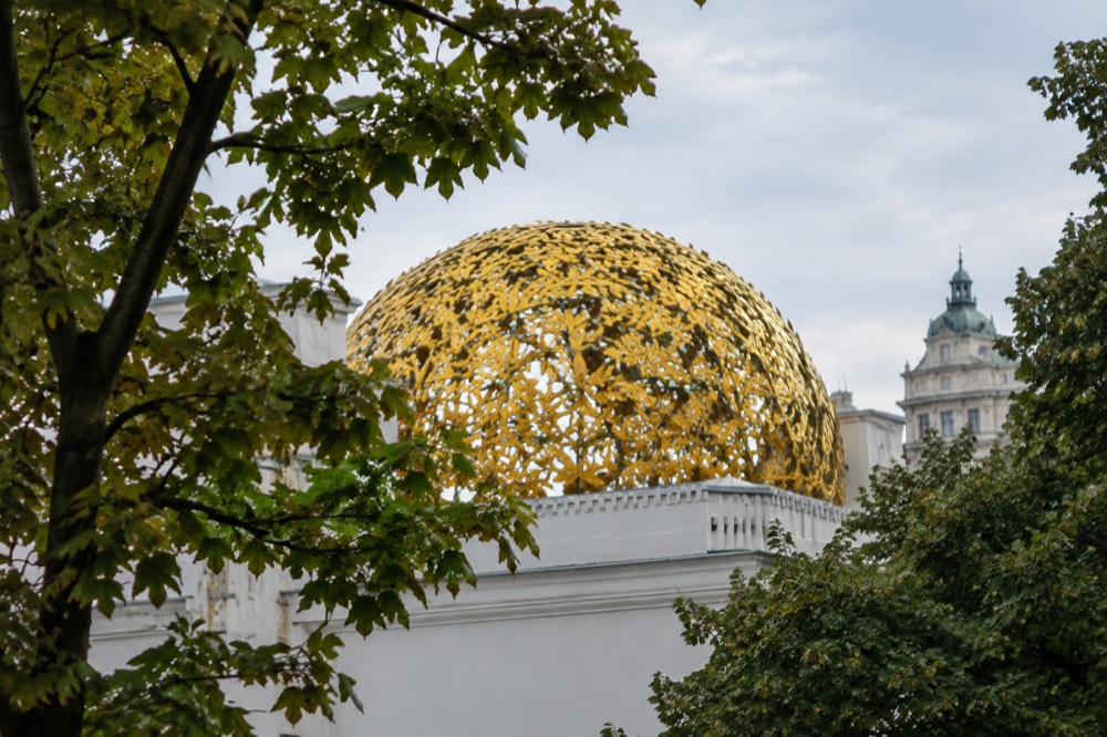 The golden openwork laurel dome of the Secession seen from below between tree branches on the Karlsplatz side, the plain white wall mostly out of frame