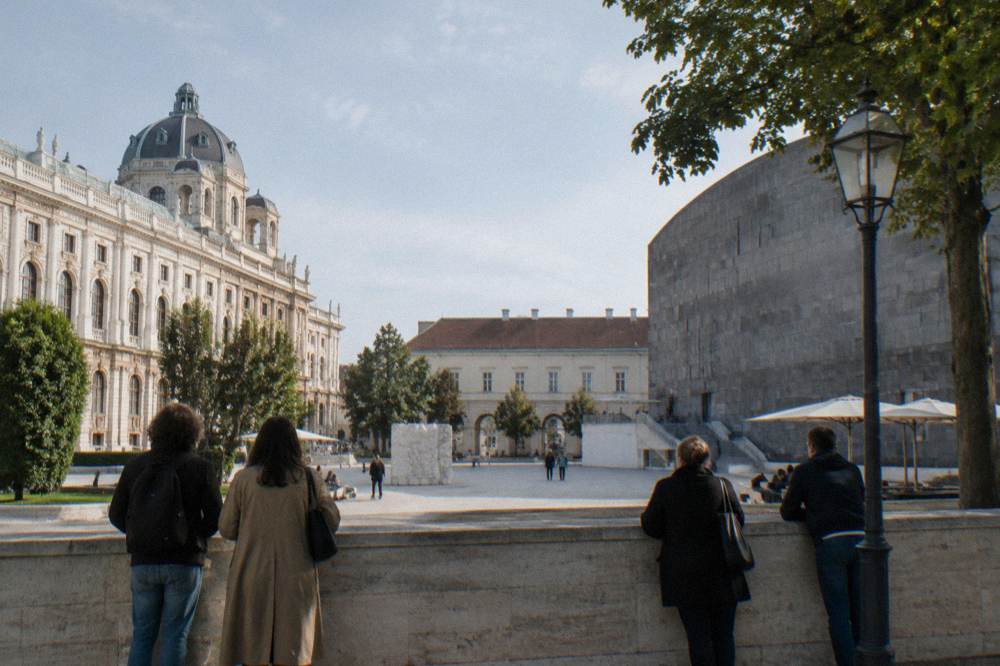 A museum forecourt on the edge of the MuseumsQuartier with a few people seen from behind waiting by a stone wall in morning light