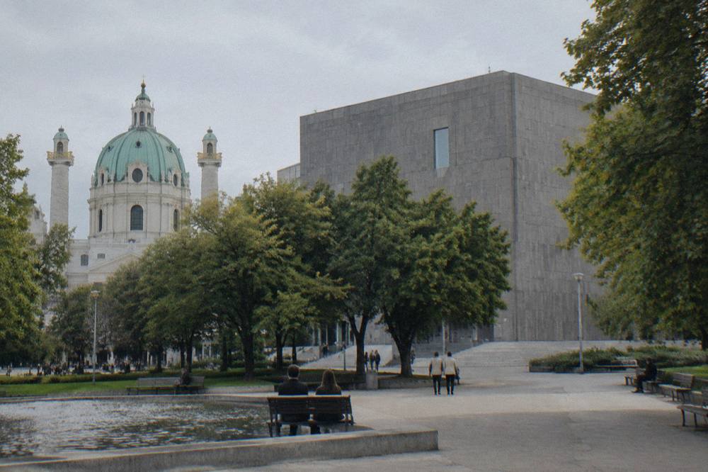 The Wien Museum's grey modern block rising behind the Karlsplatz trees, photographed from across the plaza in even light