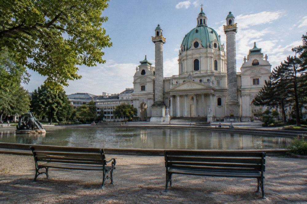 The Karlsplatz fountain pond with the Karlskirche dome rising beyond, a pair of benches empty in the foreground