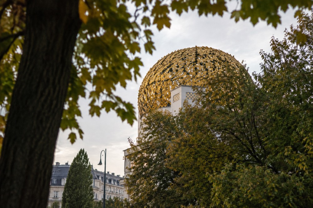The golden openwork laurel dome of the Secession seen from below between tree branches on the Karlsplatz side, the plain white wall mostly out of frame