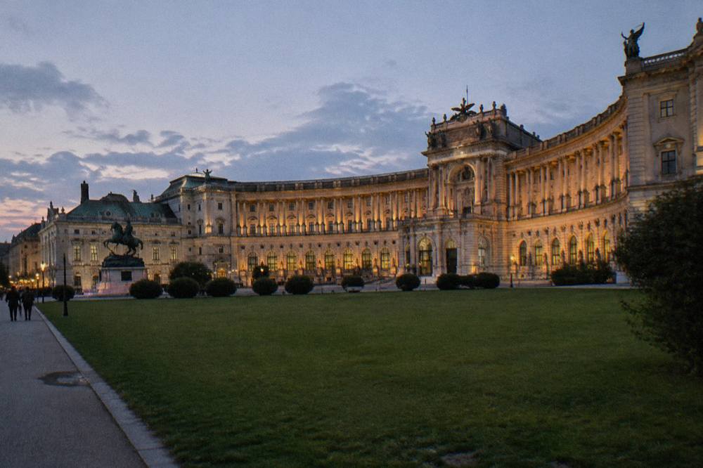 The curved Neue Burg wing on Heldenplatz at dusk with lit facades and a wide empty lawn in front