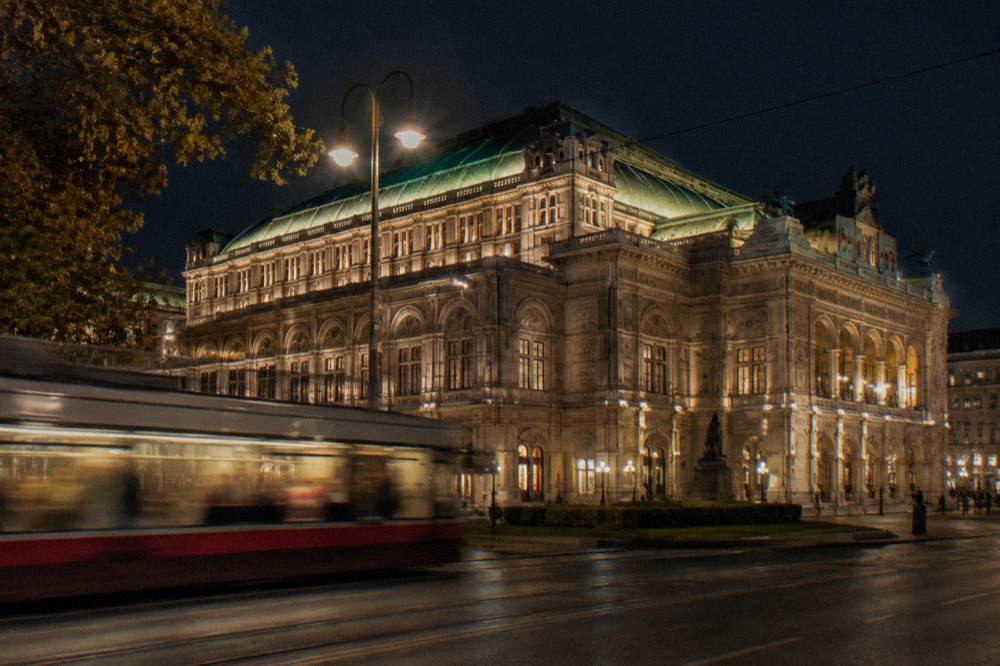 The State Opera exterior glowing on the Ringstrasse at night with a tram passing blurred in front