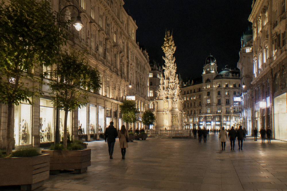 The Graben pedestrian street at night with lit shop windows and the ornate Plague Column in the middle distance