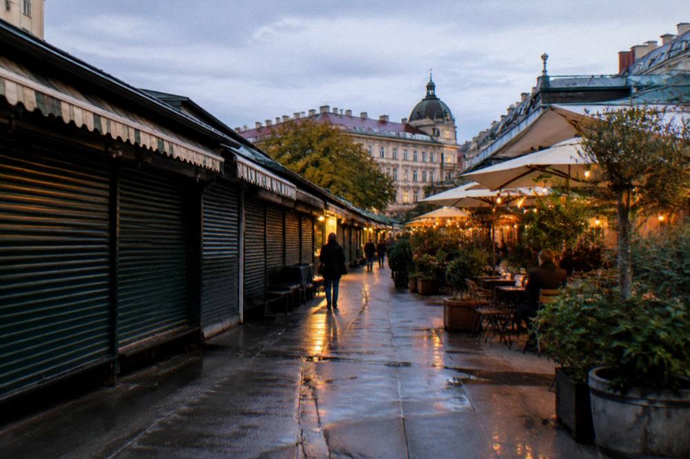 The Naschmarkt aisle in the evening with shuttered stalls, a few lit restaurant terraces and the market walkway damp underfoot
