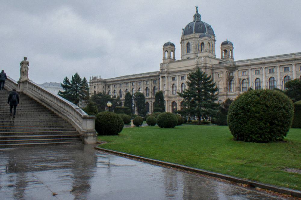 A broad staircase side of the Maria-Theresien-Platz gardens with the Naturhistorisches Museum dome rising over wet grass after rain