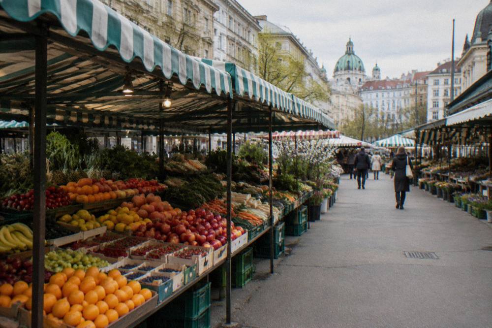 Fresh vegetable and fruit displays under striped awnings at Naschmarkt stalls, seen from the aisle with a few shoppers far off