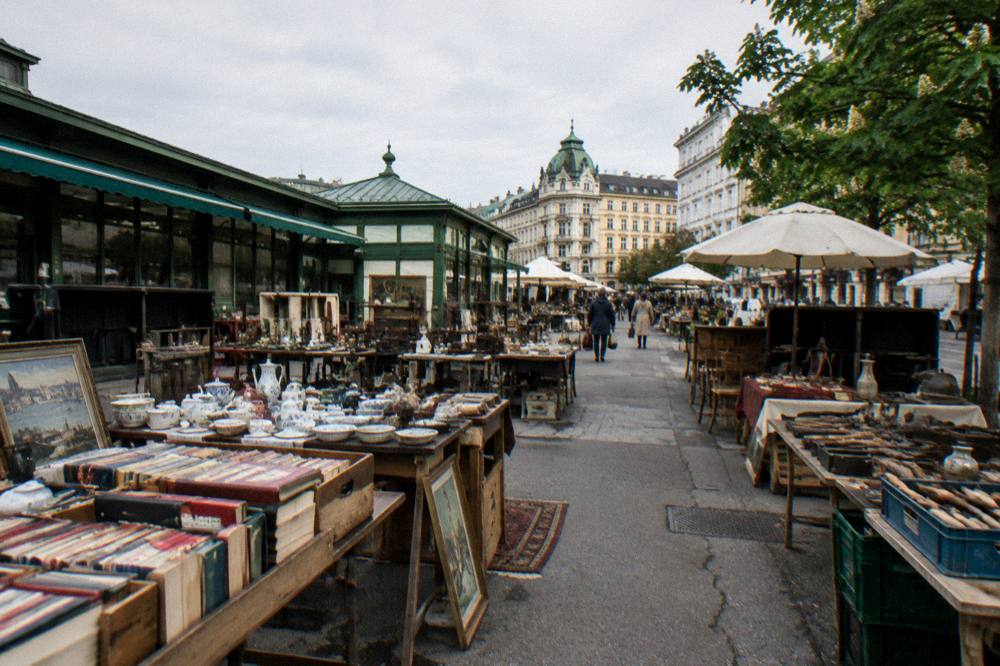 Rows of second-hand tables with books, dishes and old tools set out on the Naschmarkt flea market ground, buyers tiny in the distance