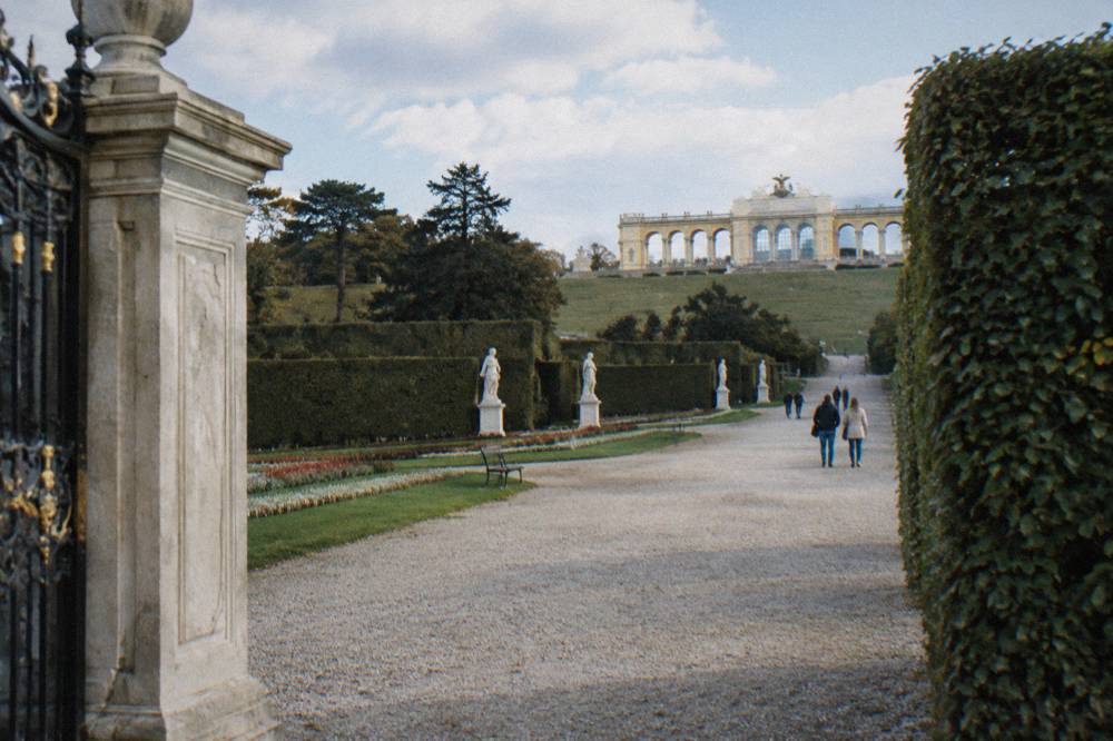 A stone gate and trimmed hedge framing a gravel garden path that leads uphill toward the Gloriette colonnade