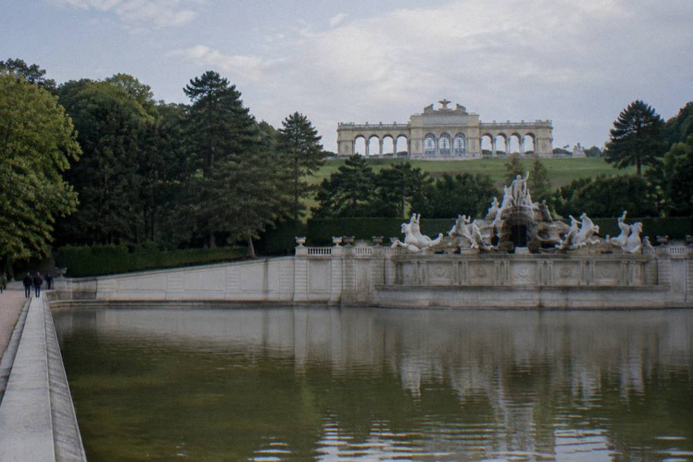 The Neptune Fountain basin with its sculpted stone figures below the Gloriette hill, water still and trees behind