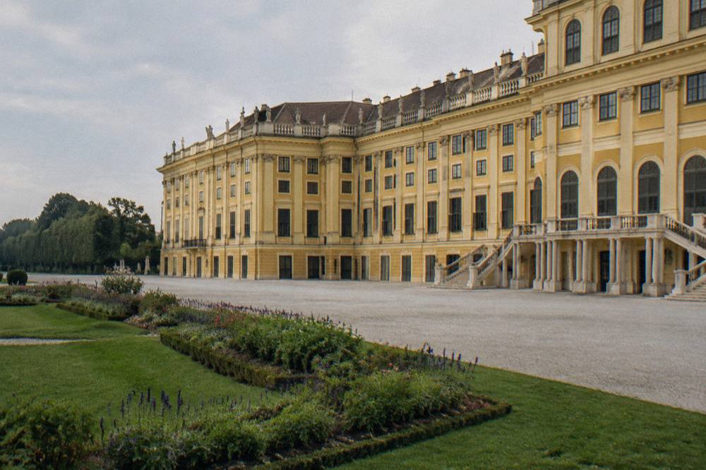 The ochre yellow Schönbrunn facade seen from the edge of the gravel parterre side with planted borders and no visitors in frame