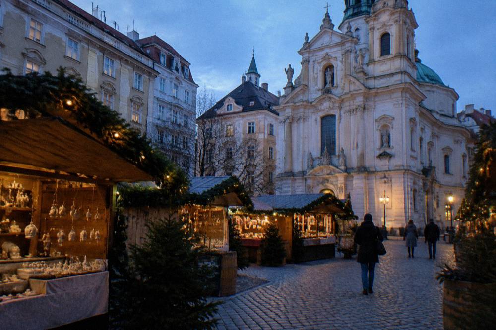 Wooden stall roofs hung with evergreen garlands on the Am Hof cobbles, the pale Baroque church facade behind in blue evening light
