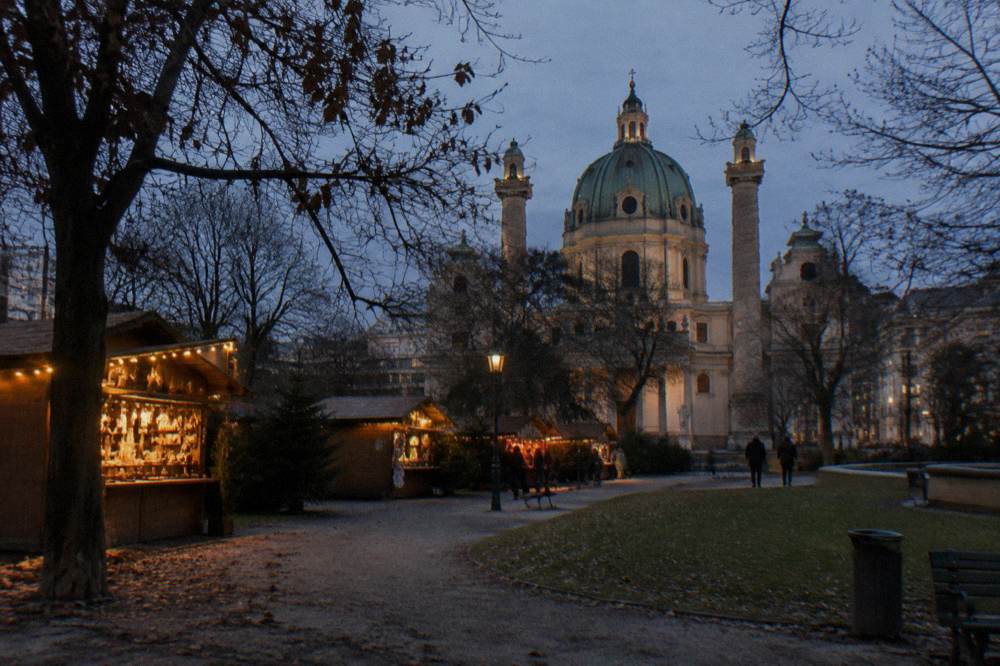 Bare trees and warm lights on small wooden stalls in the Resselpark at dusk with the Karlskirche dome dark behind