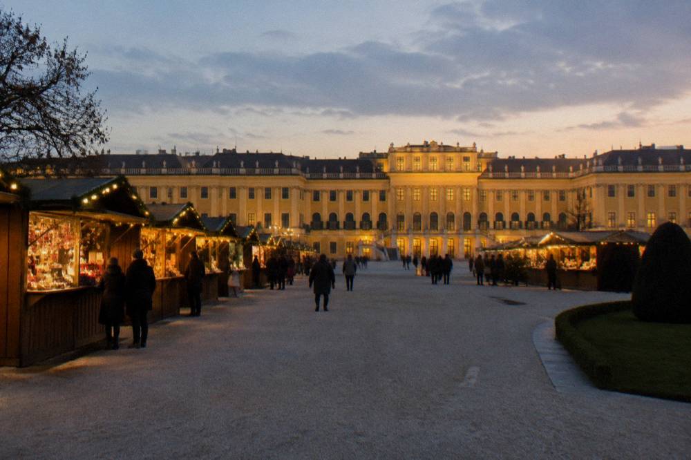 The forecourt of Schönbrunn Palace at dusk with rows of lit wooden stall roofs and the yellow facade glowing beyond
