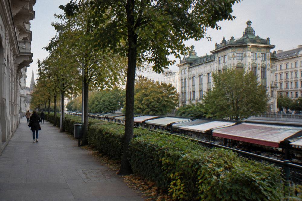 The Linke Wienzeile pavement beside the Naschmarkt with the Otto Wagner majolica house facade beyond the tree line