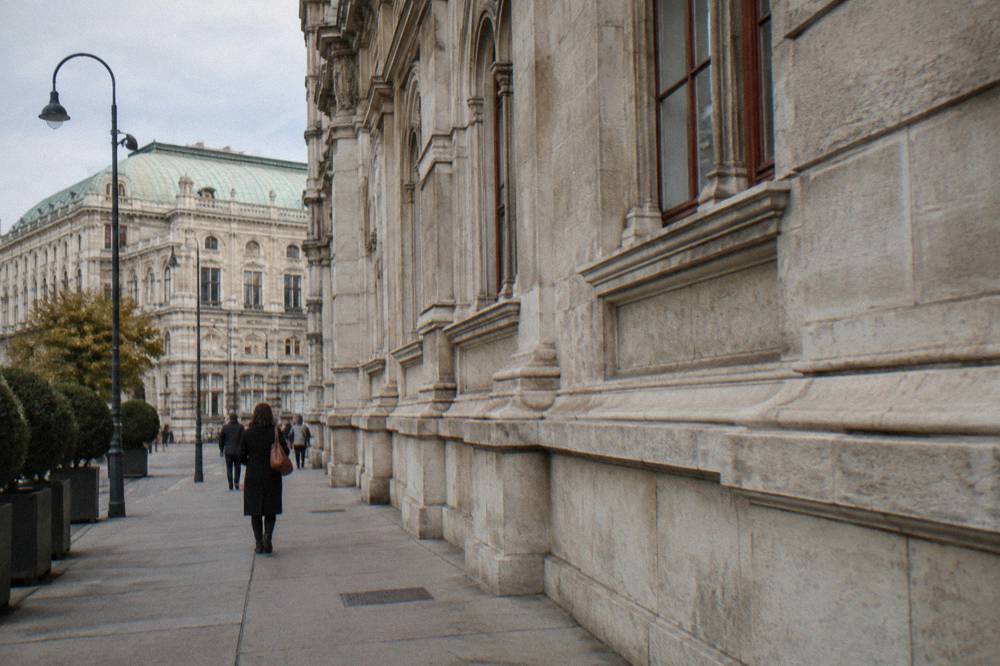 The side wall of the State Opera on Operngasse, plain stonework and a pavement with a few distant passers-by from behind