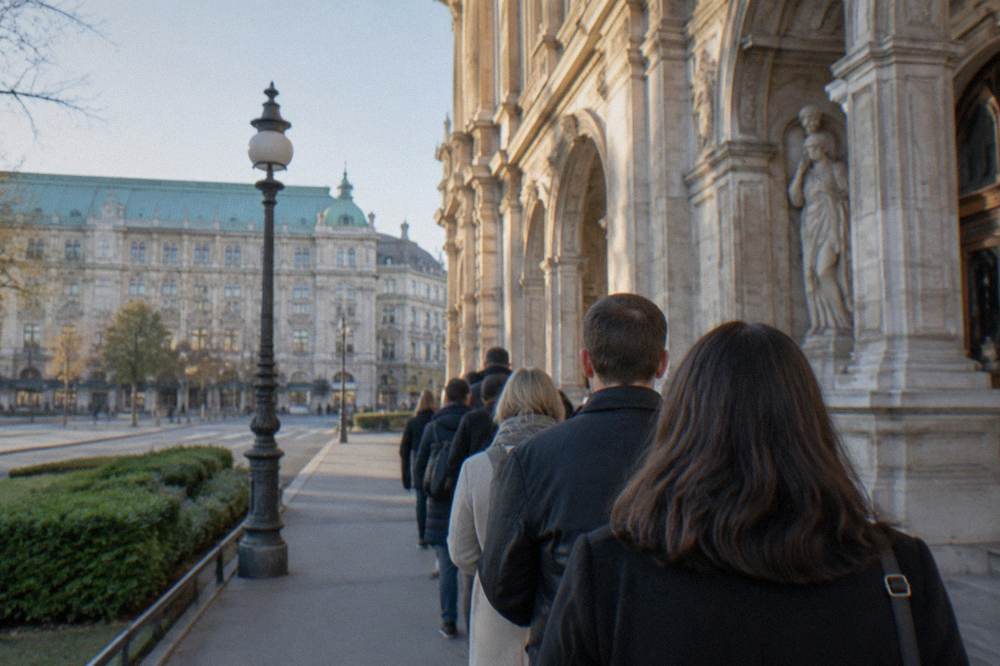 A line of people seen from behind waiting on the pavement beside the State Opera wall in morning light